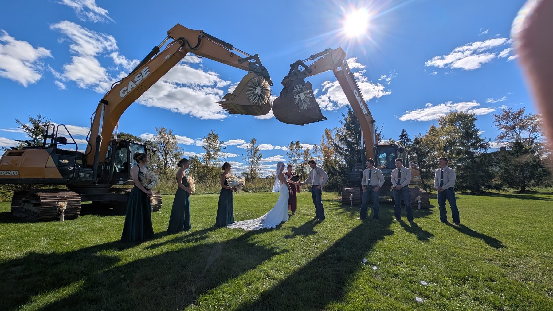A wedding party gathered on a lawn under decorated excavators as Tammy officiates
